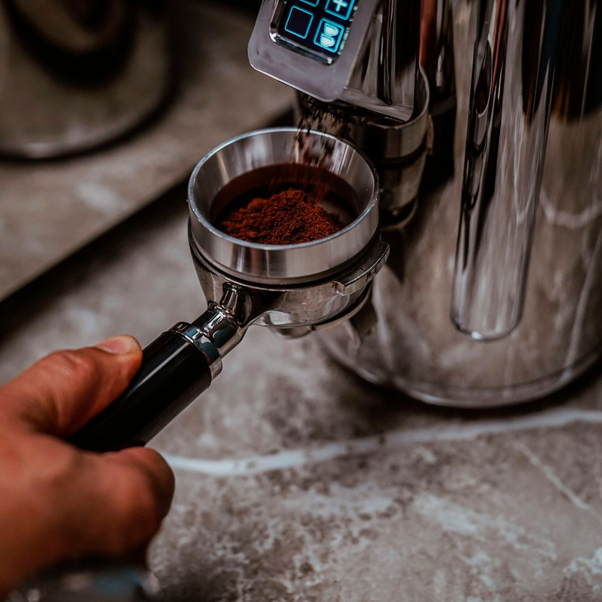 Eine Hand hält ein Siebträger, während dunkle Kaffeepulver von einer Mühle hineinfällt.