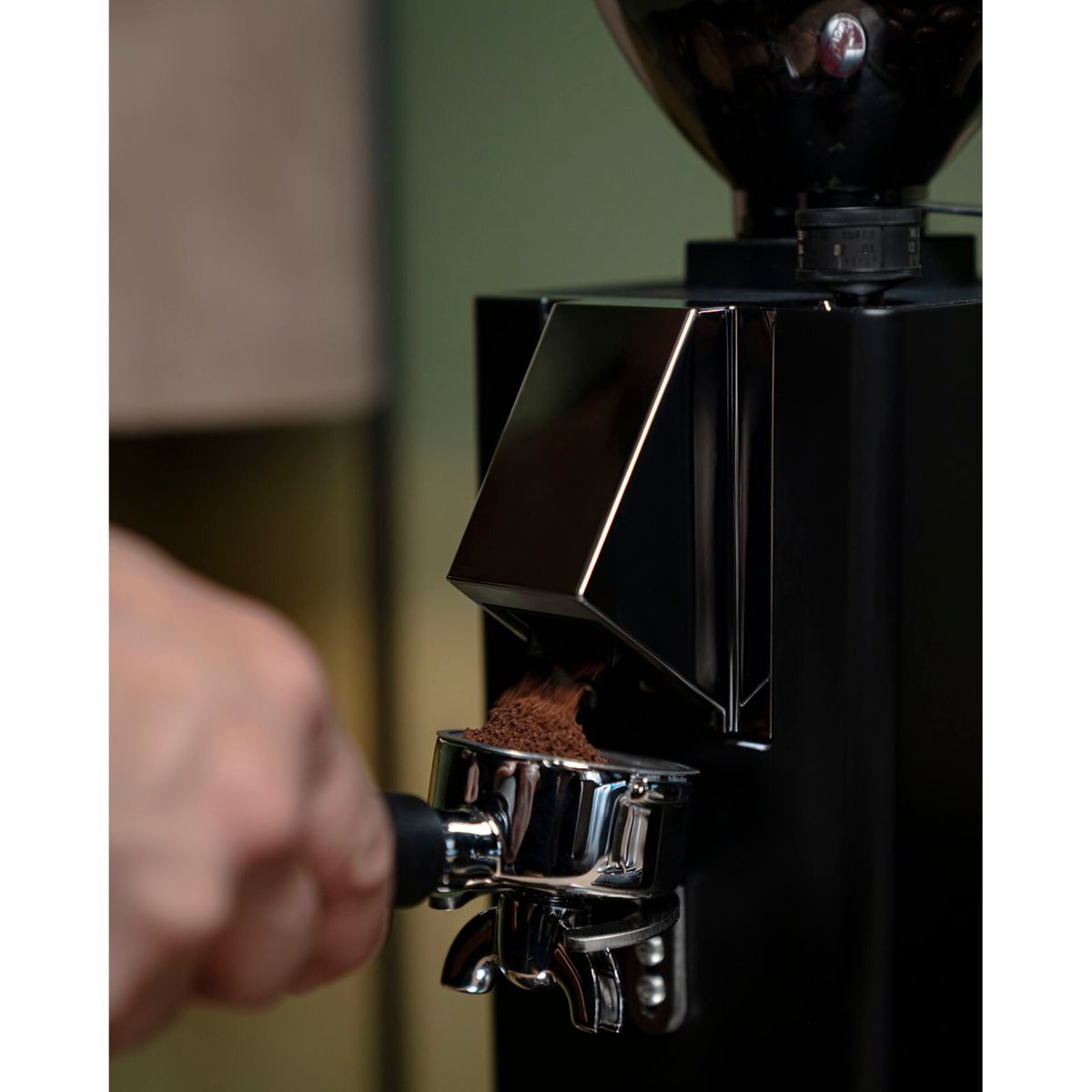 Una mano tiene un portafiltro sotto un macinacaffè nero mentre cade caffè macinato.