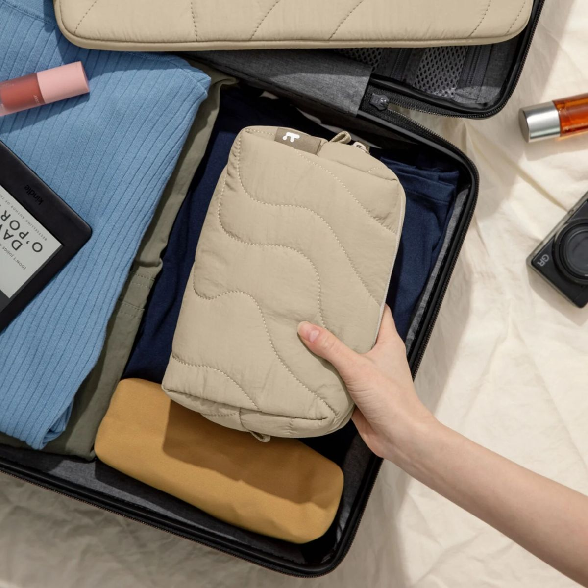 Eine Hand legt eine beige gesteppte Tasche in einen offenen Koffer voller Kleidung.