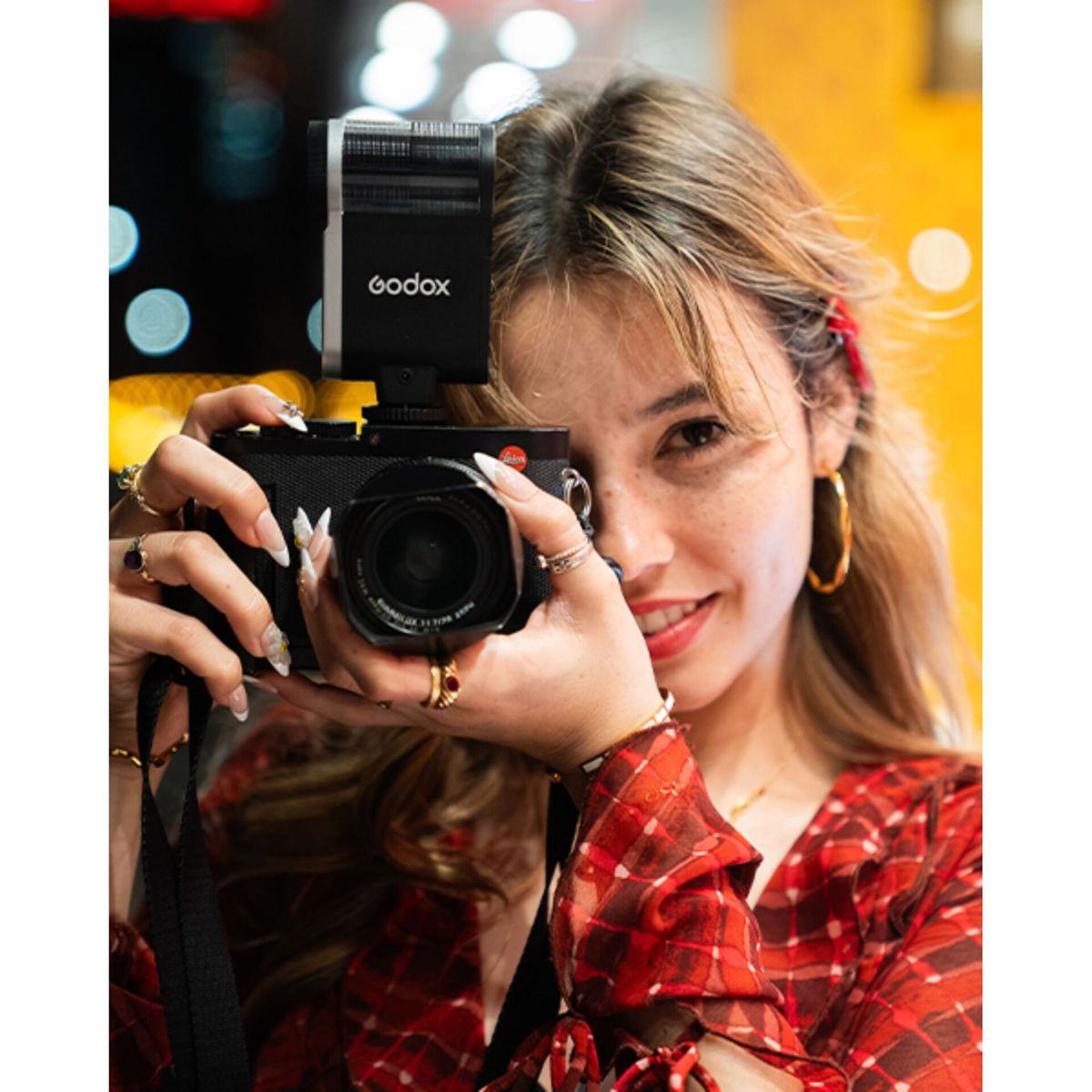 Una donna con capelli lunghi e abiti a quadri tiene una fotocamera con flash.