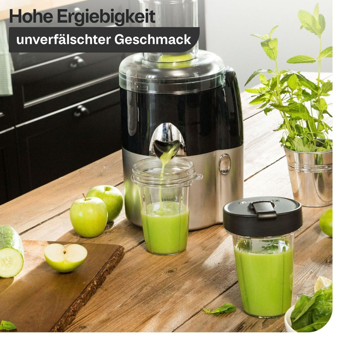 Ein Entsafter presst grünen Saft in Becher. Äpfel und Gurke liegen daneben.