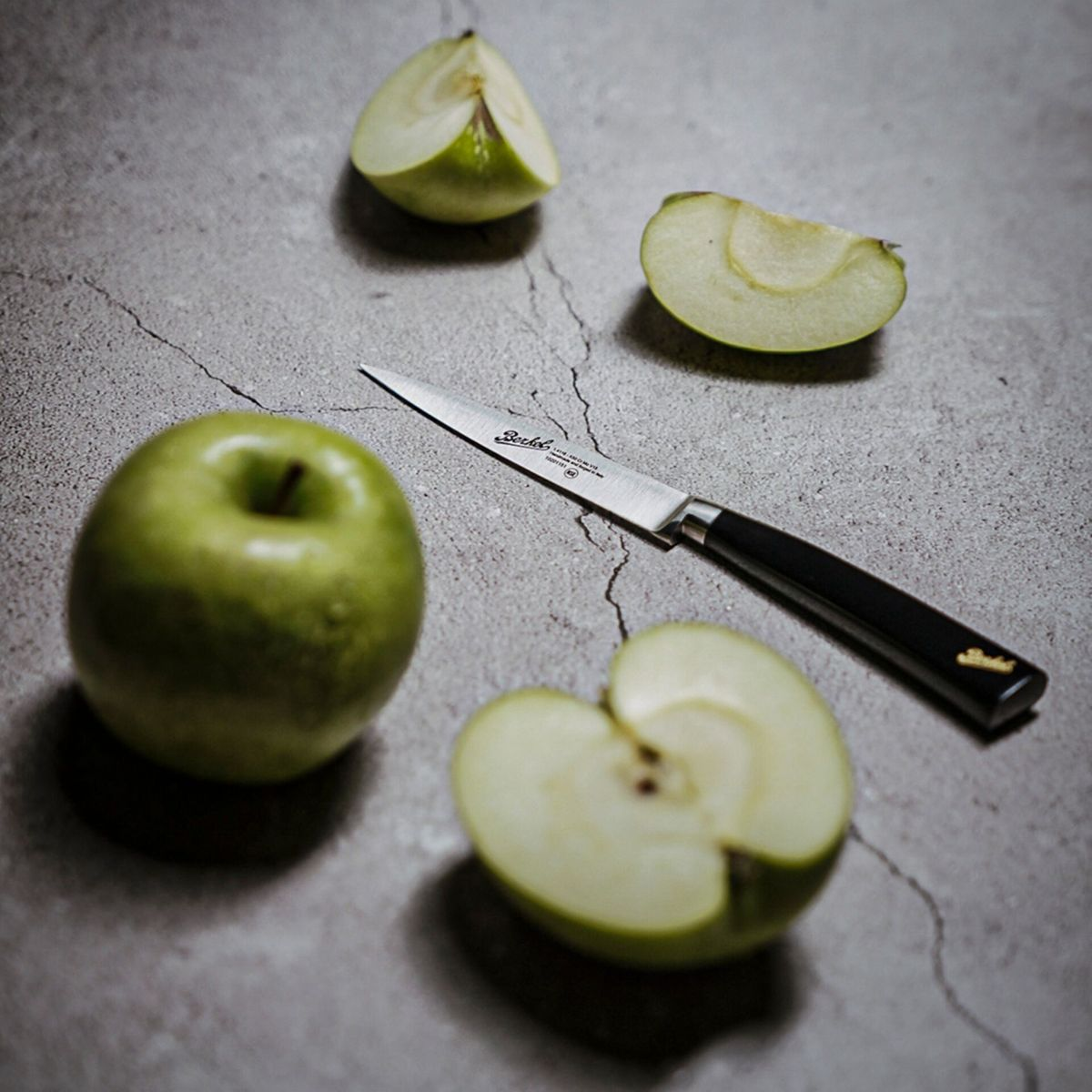 Ein Messer liegt auf einer grauen Oberfläche neben einem ganzen grünen Apfel und Apfelscheiben.