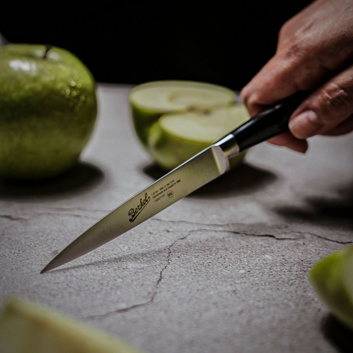 Eine Hand hält ein Messer, das einen grünen Apfel auf einer Steinplatte schneidet. Andere Äpfel sind sichtbar.