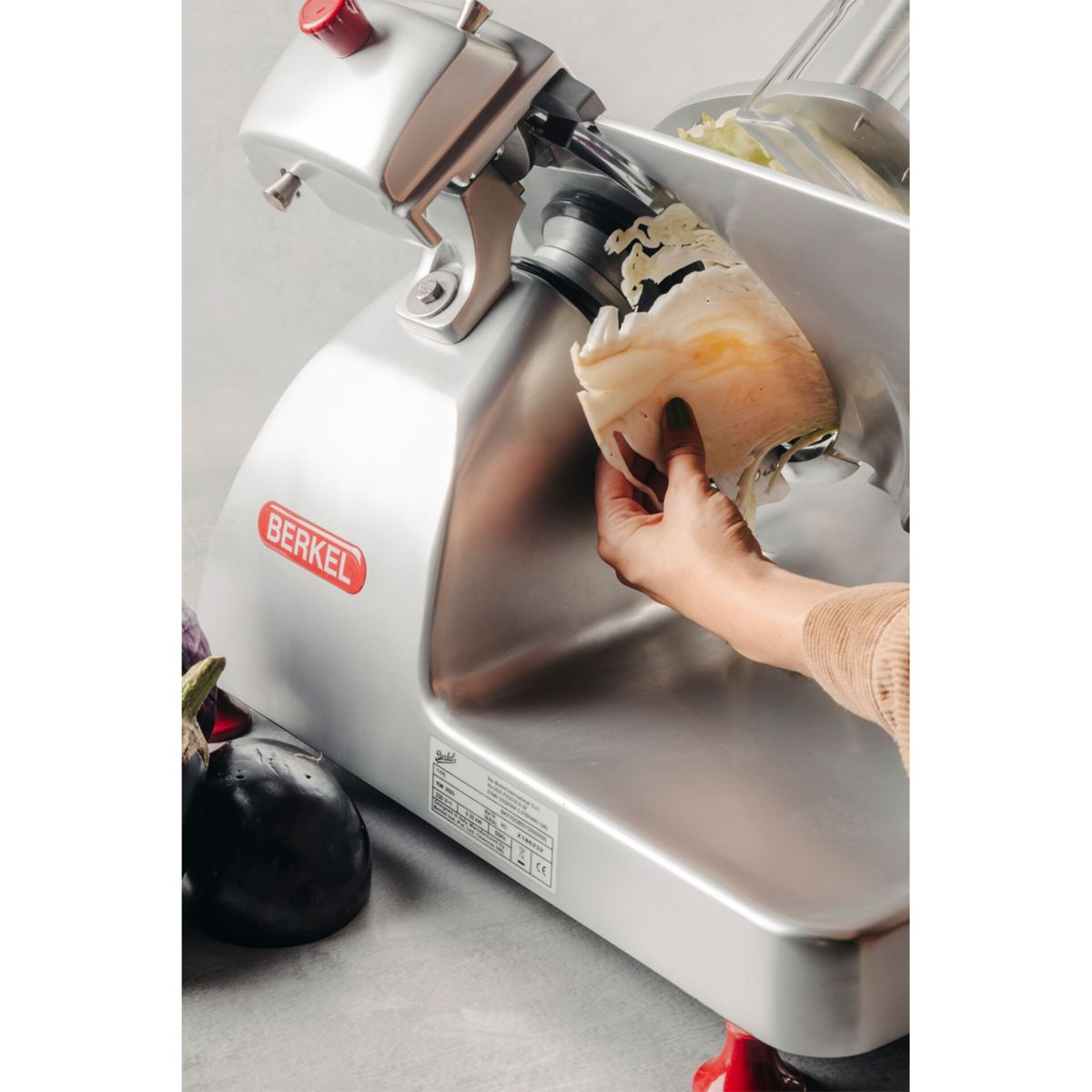 Eine Hand hält Käse auf einer Berkel-Schneidemaschine neben Auberginen.