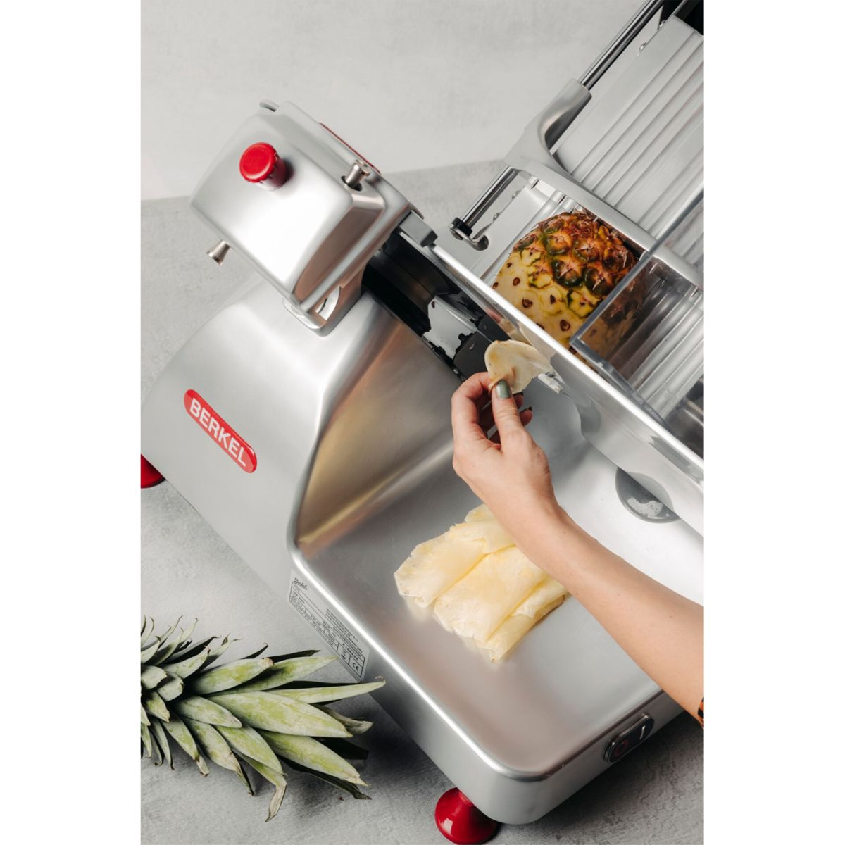 Eine Hand legt Ananasscheiben auf eine Silbermaschine. Unten liegt eine Ananas.