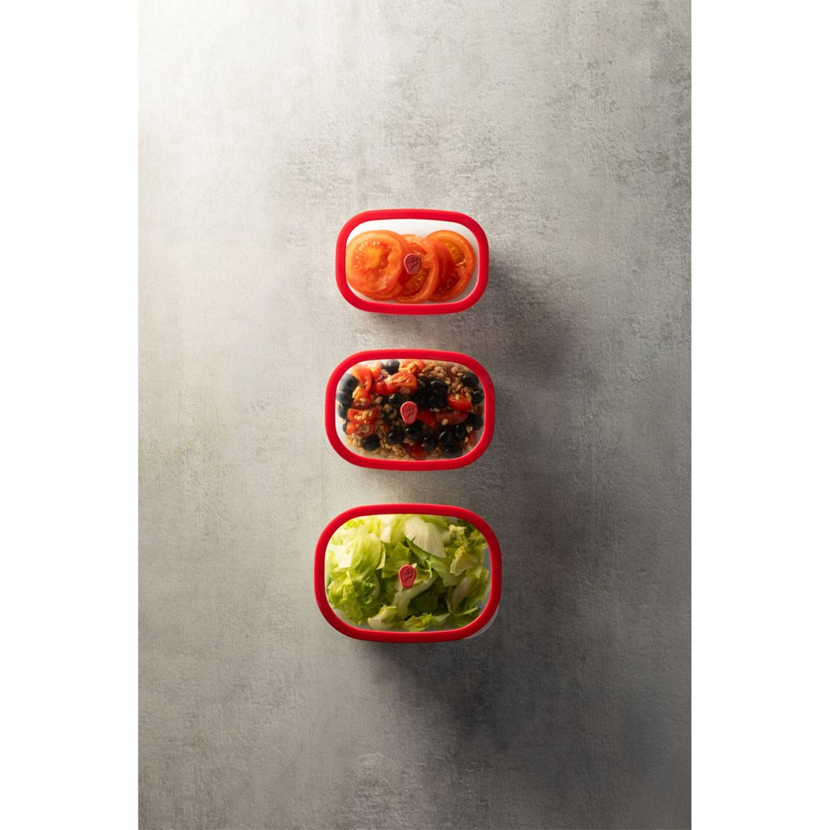 Drei rote Behälter mit Tomaten, Salat und Getreide auf grauem Hintergrund.