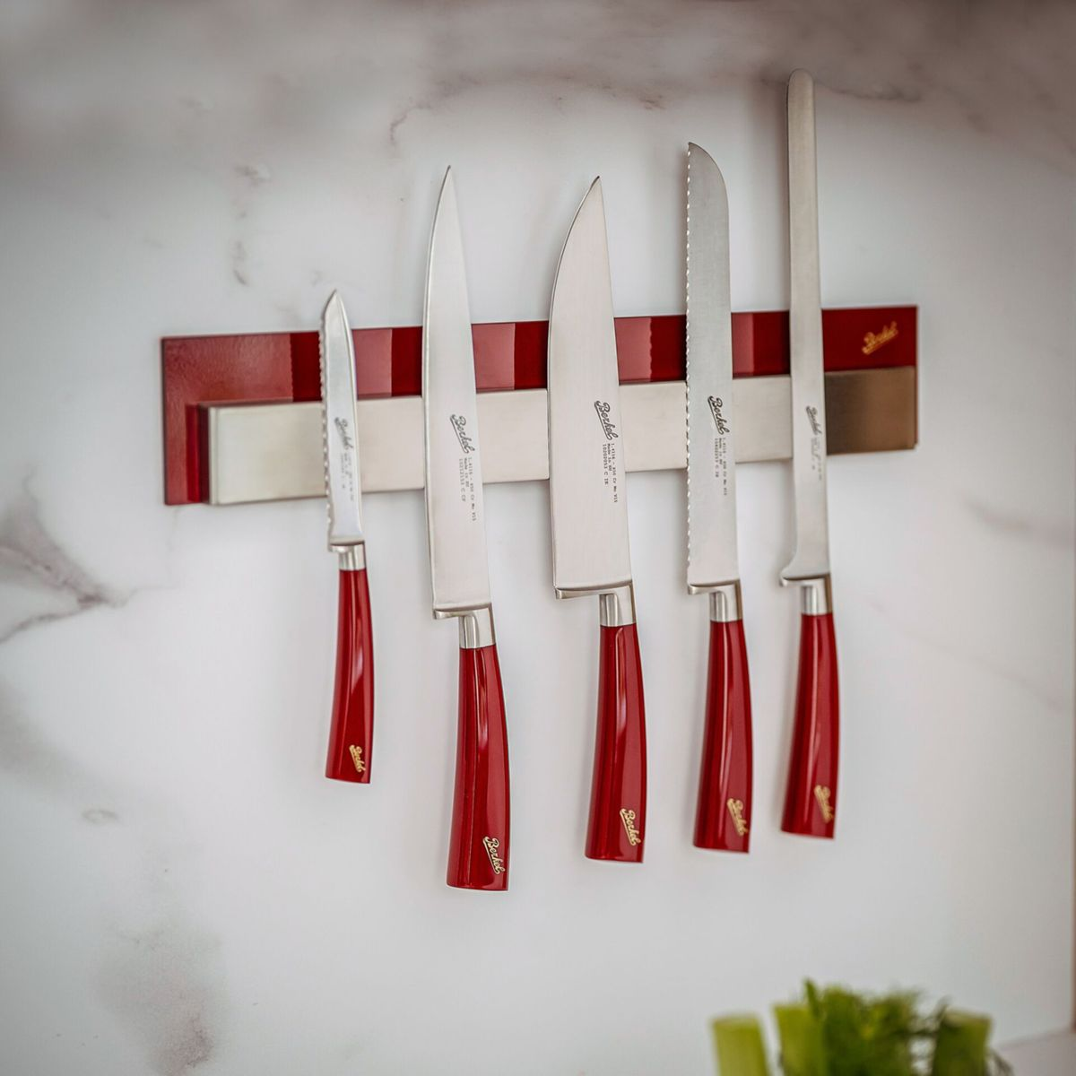 Fünf Messer mit roten Griffen hängen an einer magnetischen Leiste an einer Marmorwand.