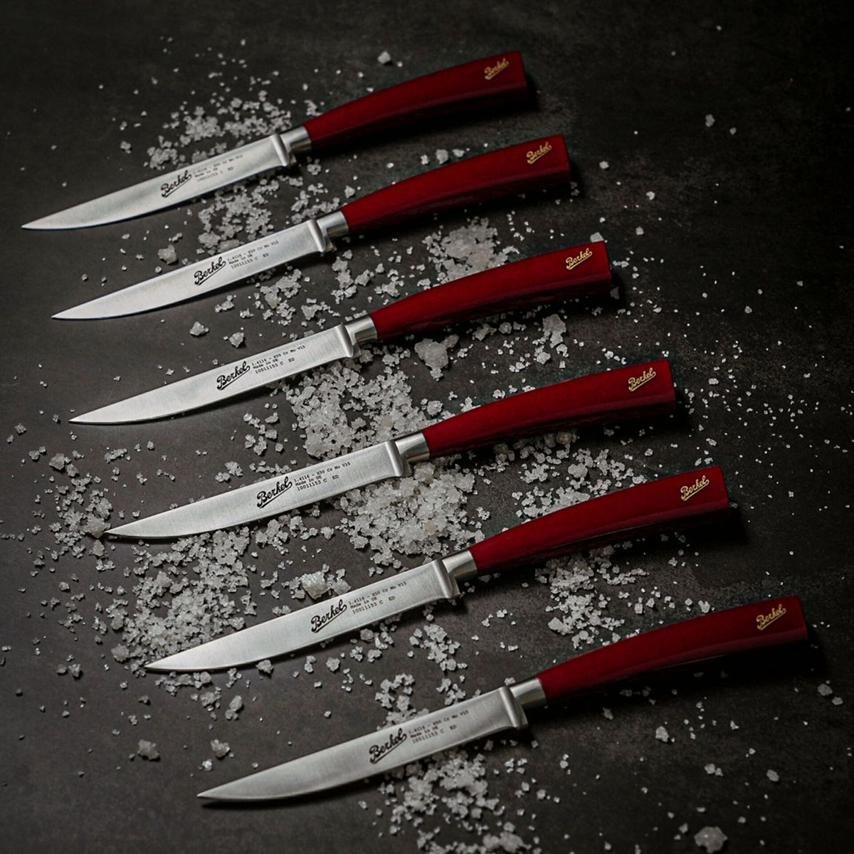 Sechs Messer mit roten Griffen und silbernen Klingen liegen auf Salz.