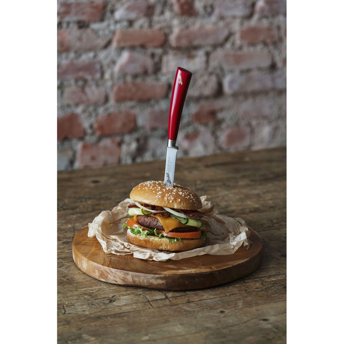 Ein Burger mit rotem Messer im Brötchen auf einem Holzteller vor Backsteinwand.