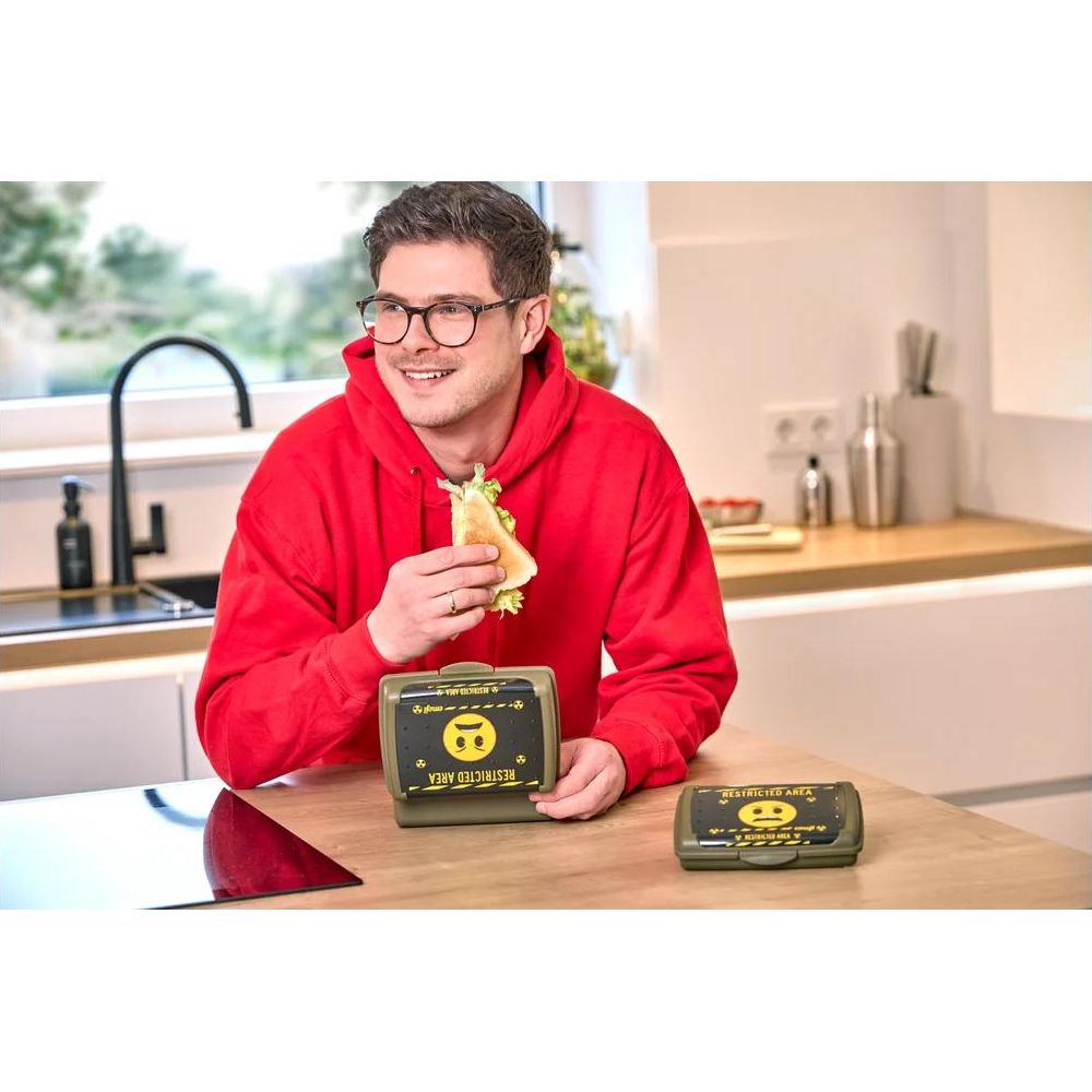 Mann in rotem Hoodie isst Sandwich neben Brotdosen mit Smiley-Motiv.