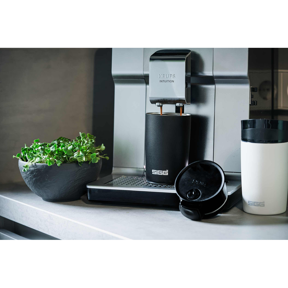 Kaffeemaschine brüht Kaffee in schwarzen Becher neben grüner Pflanze und weißem Becher.