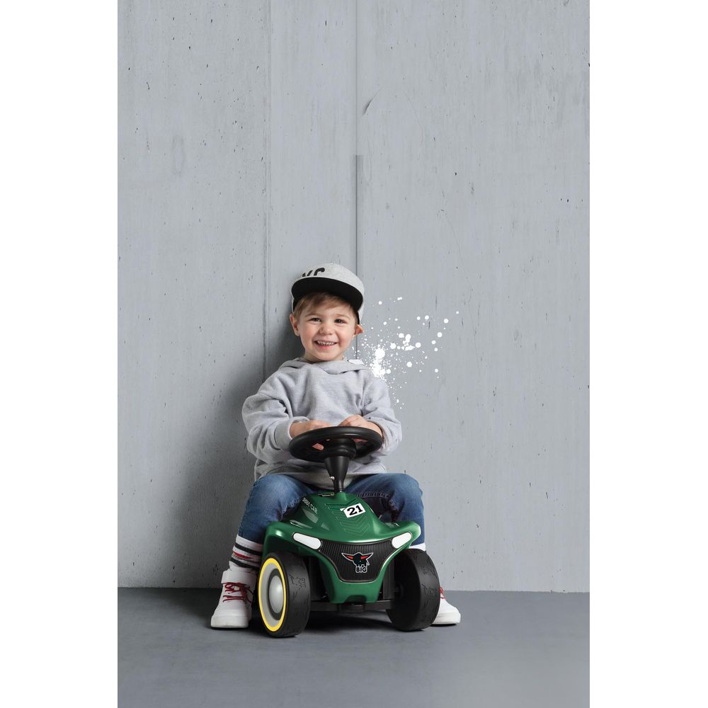 Ein Junge mit grauer Kapuzenjacke und Cap sitzt auf einem grünen Spielzeugauto.