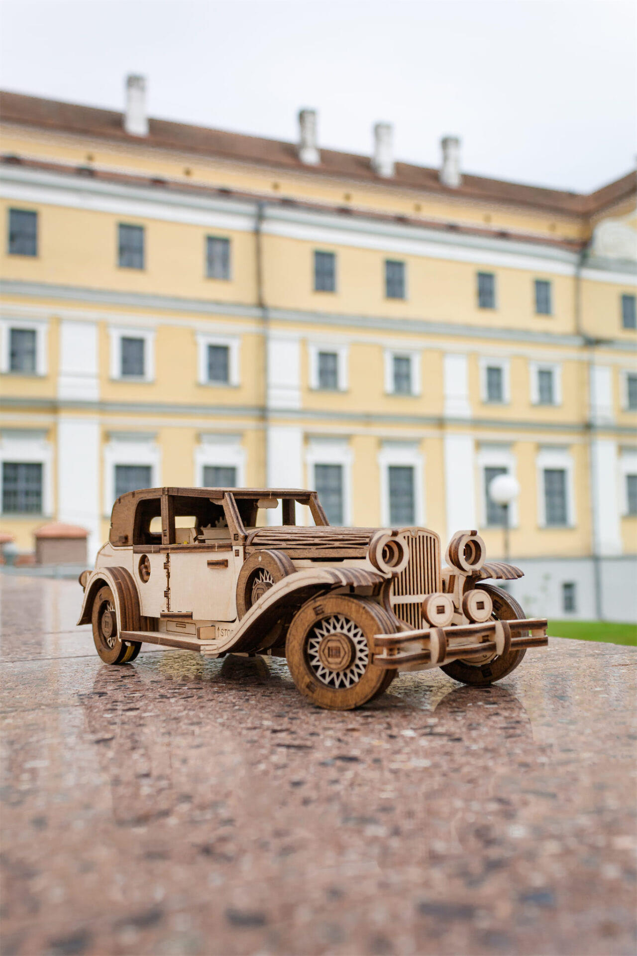 Ein Holzmodell eines Oldtimer-Autos steht auf einer Steinplatte vor einem gelben Gebäude.