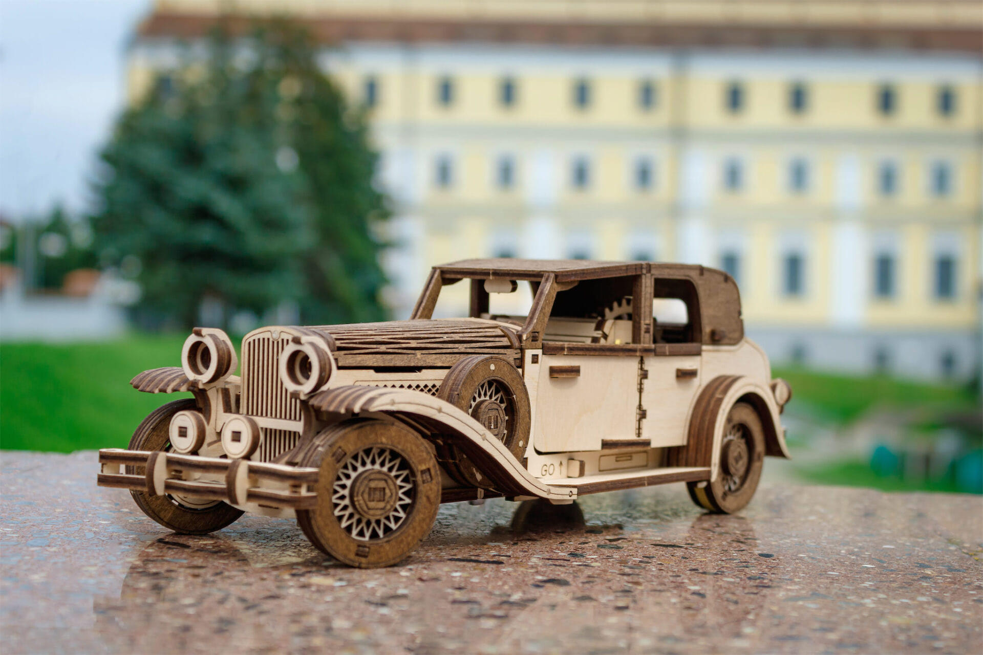 Ein hölzernes Modellauto im Vintage-Stil steht auf einer Marmorfläche vor einem Gebäude.