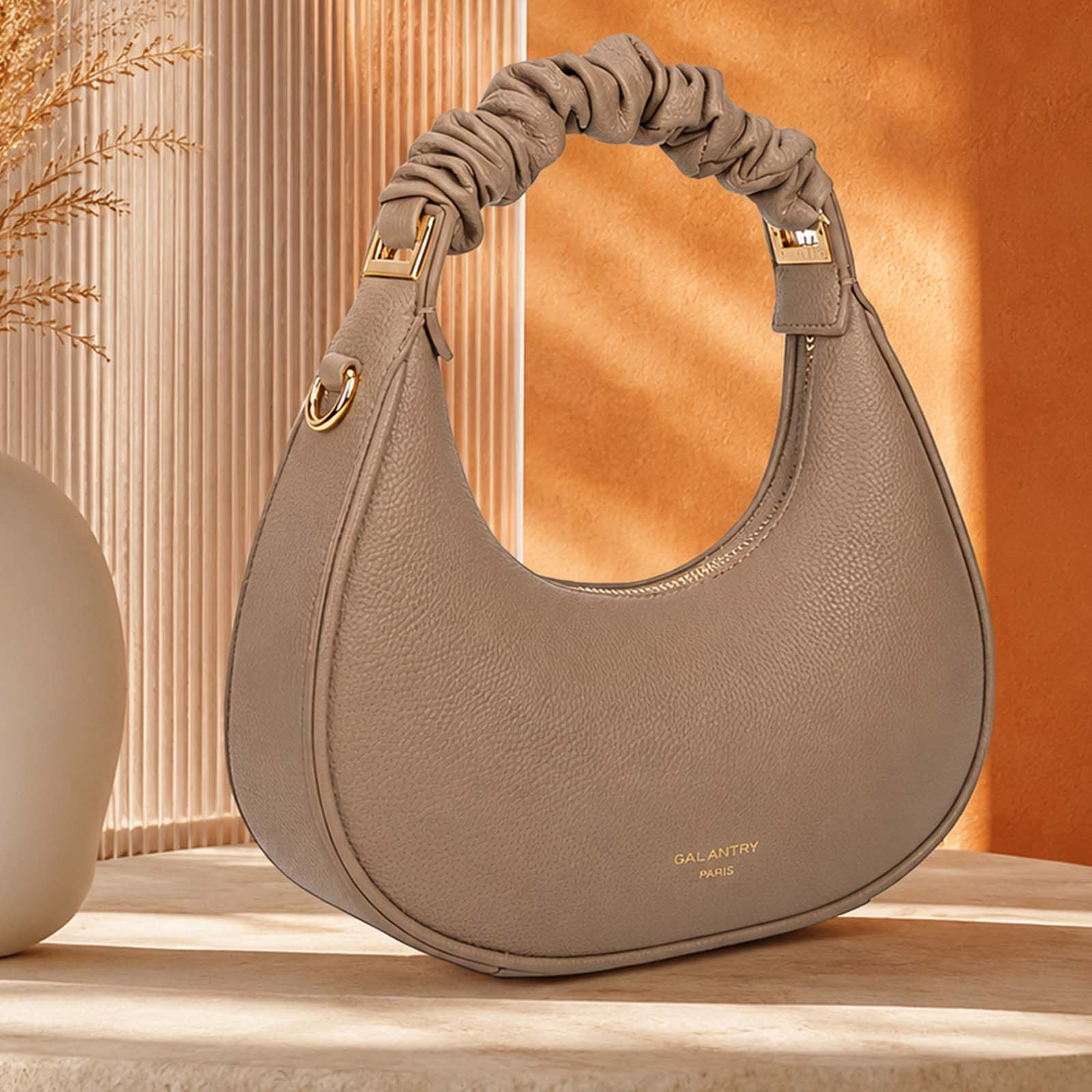 Un bolso taupe con asa fruncida y detalles dorados descansa sobre una mesa.