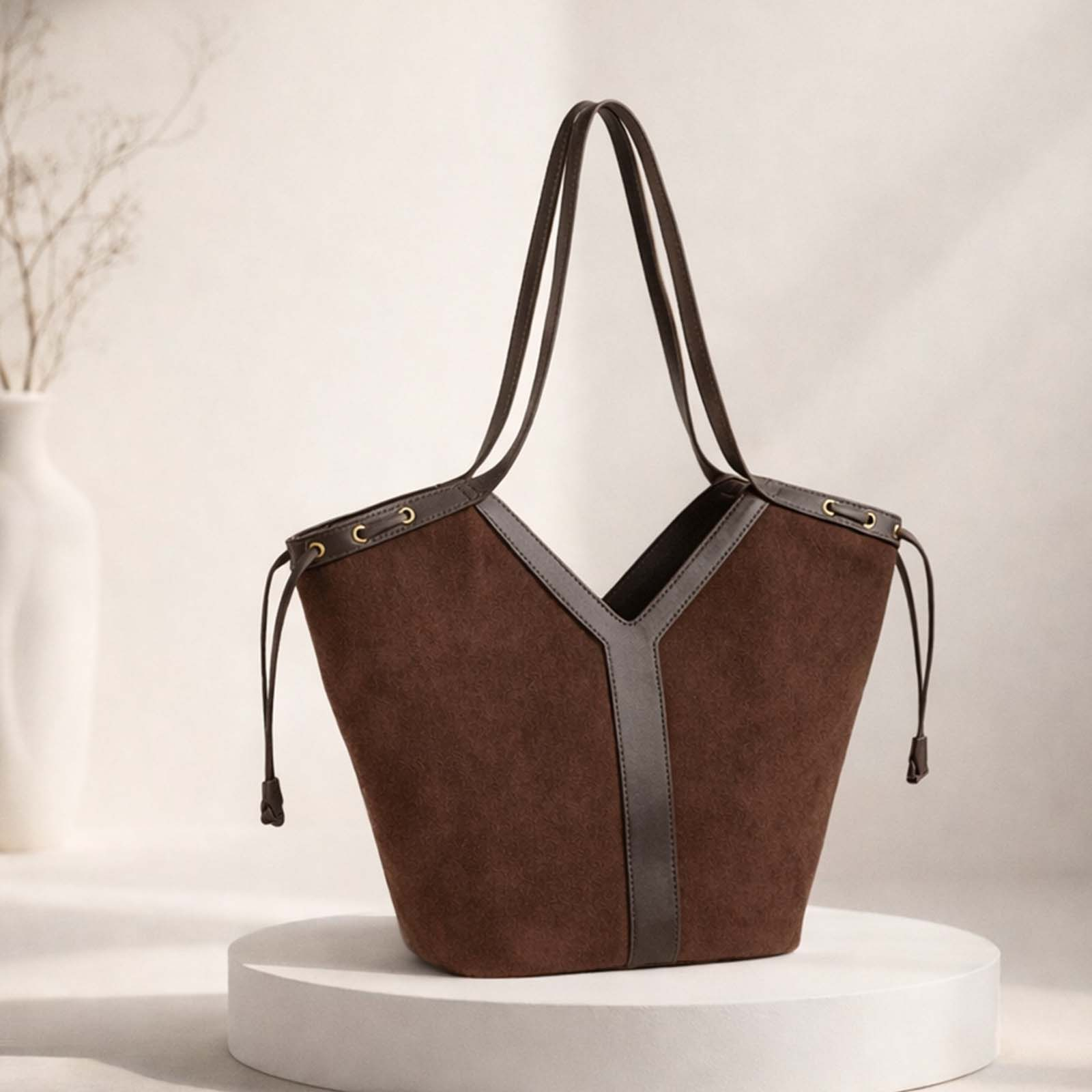 Bolso tote de ante marrón con ribete y correas de cuero oscuro, sobre un pedestal blanco.