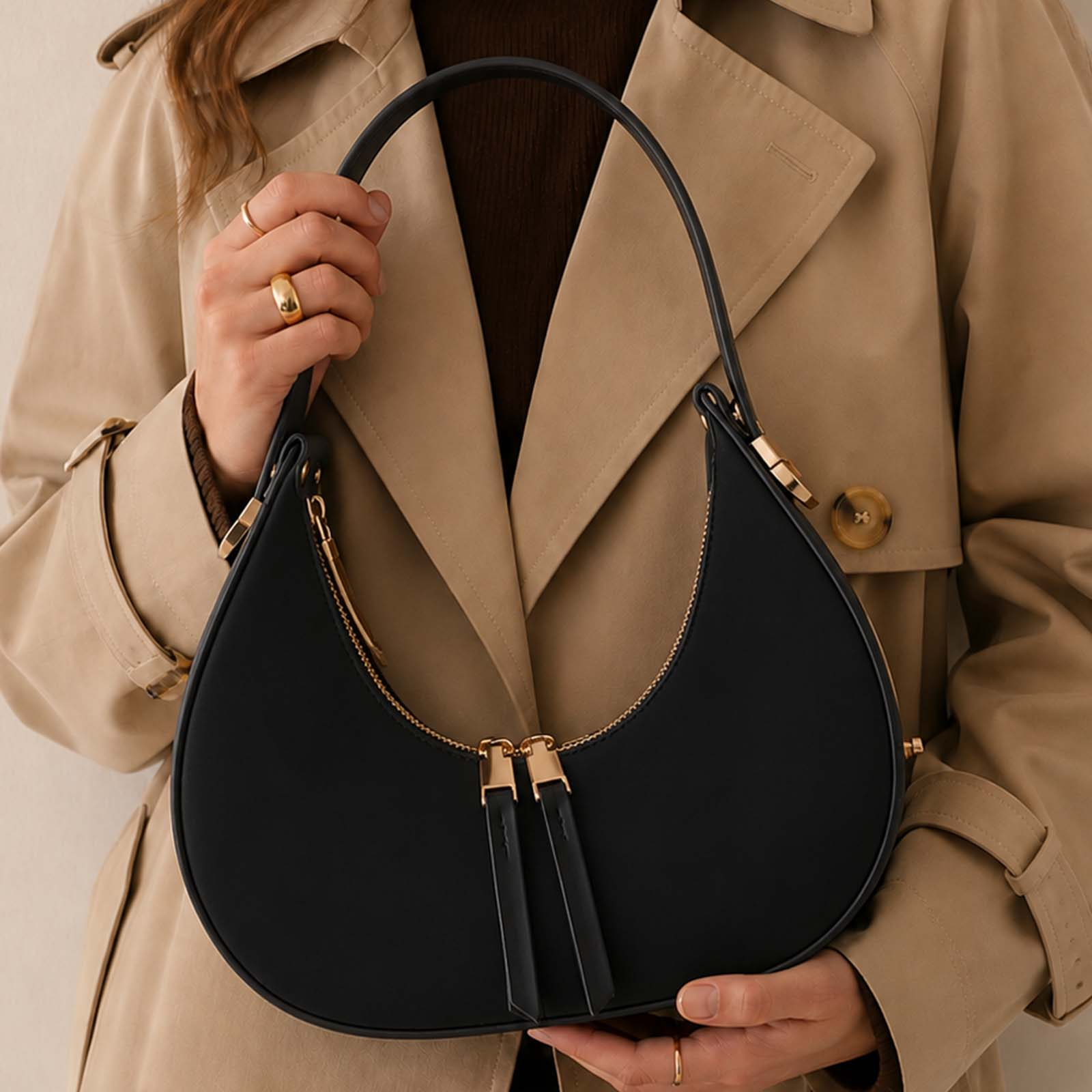 Una mujer con un abrigo beige sostiene un bolso negro de media luna con cremallera dorada.