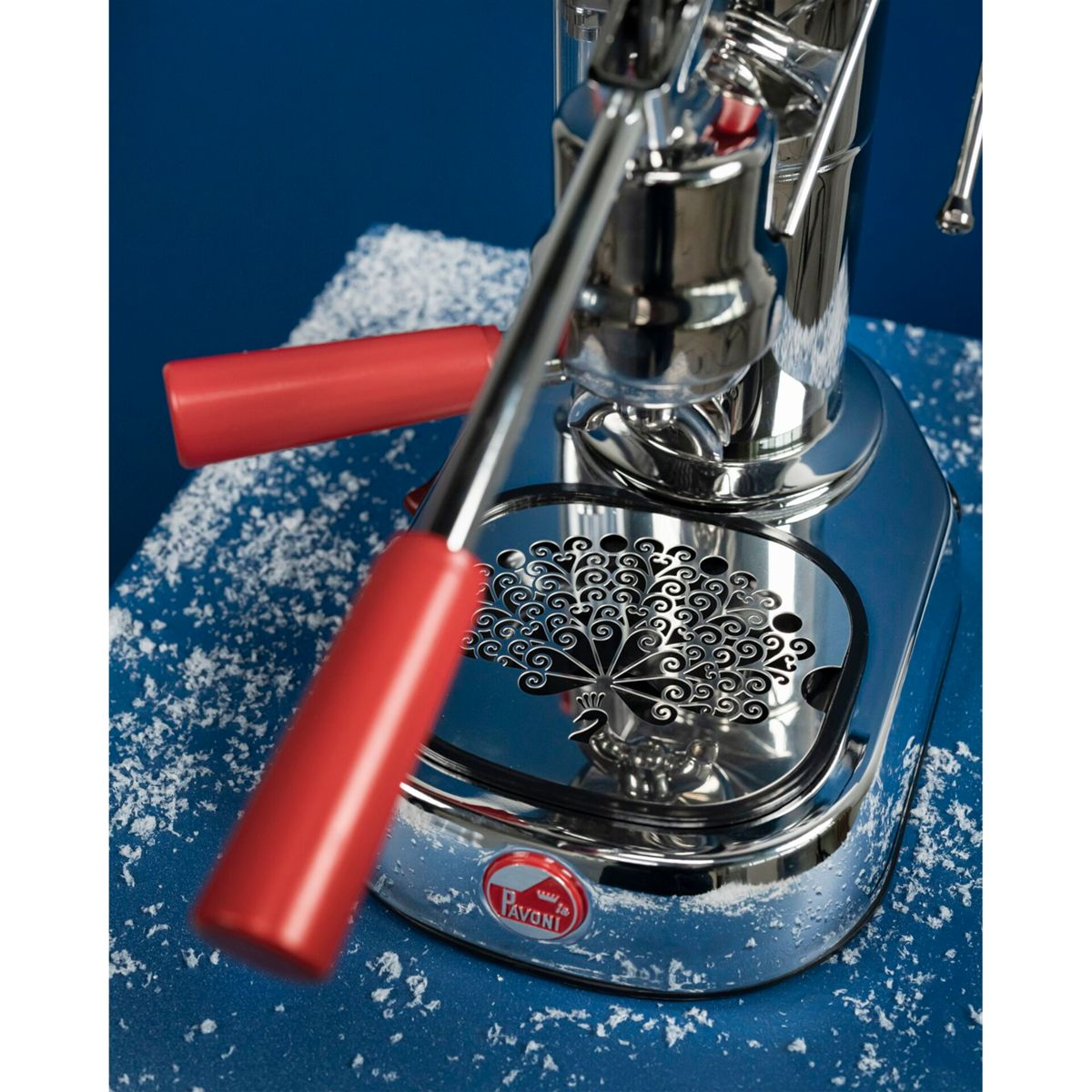 Chrom-Espressomaschine mit roten Hebeln und Pfauenmotiv auf blauem Hintergrund.