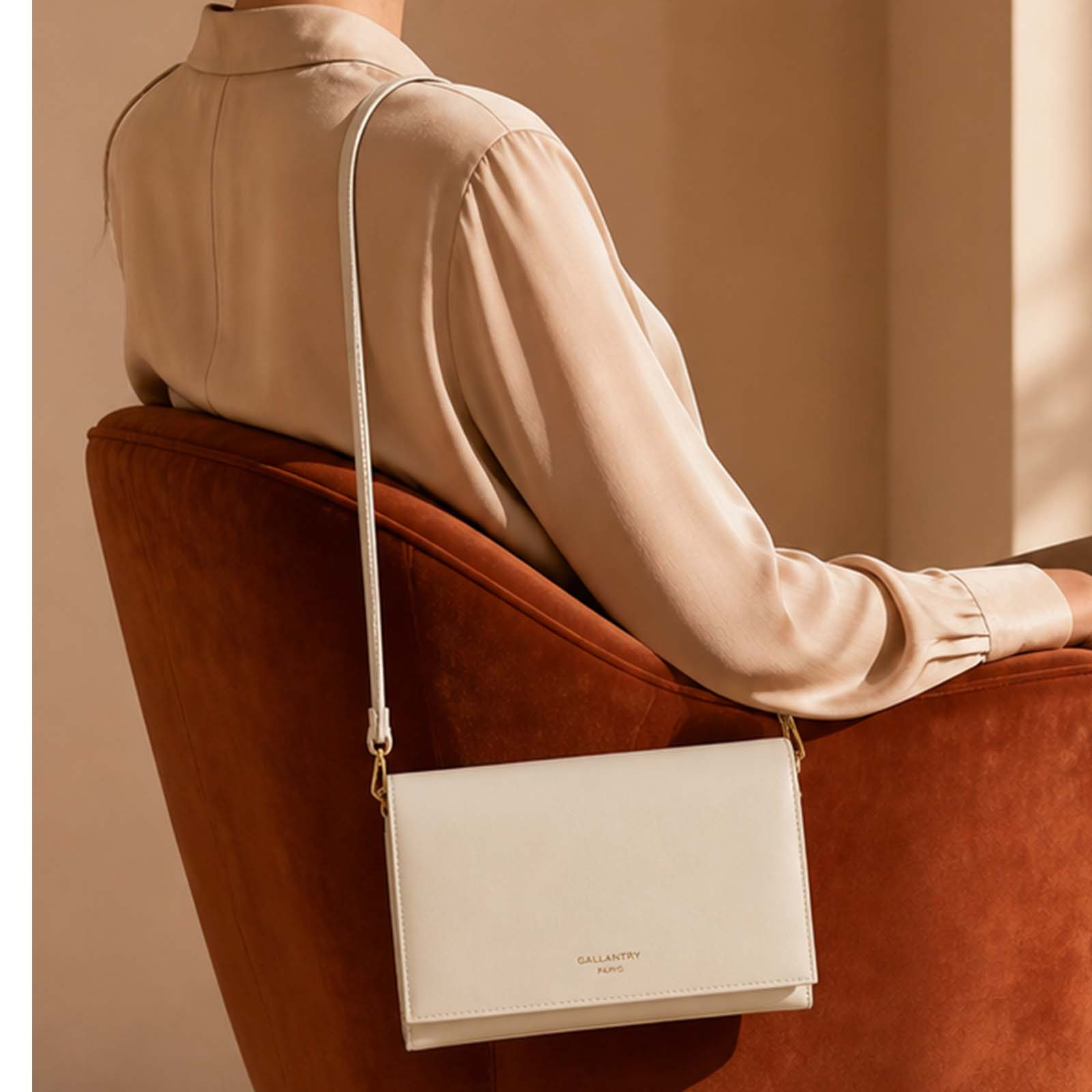 Una persona con blusa beige se sienta en silla color óxido, sosteniendo un bolso crema.