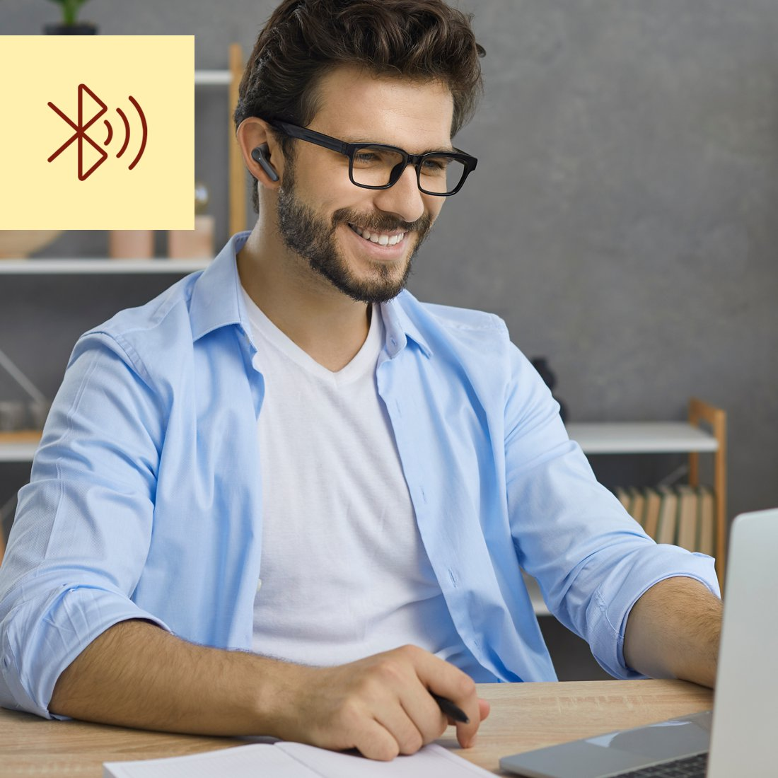 Ein Mann mit Brille und Ohrhörer lächelt, während er auf einen Laptop schaut, mit einem Bluetooth-Symbol in der Ecke.