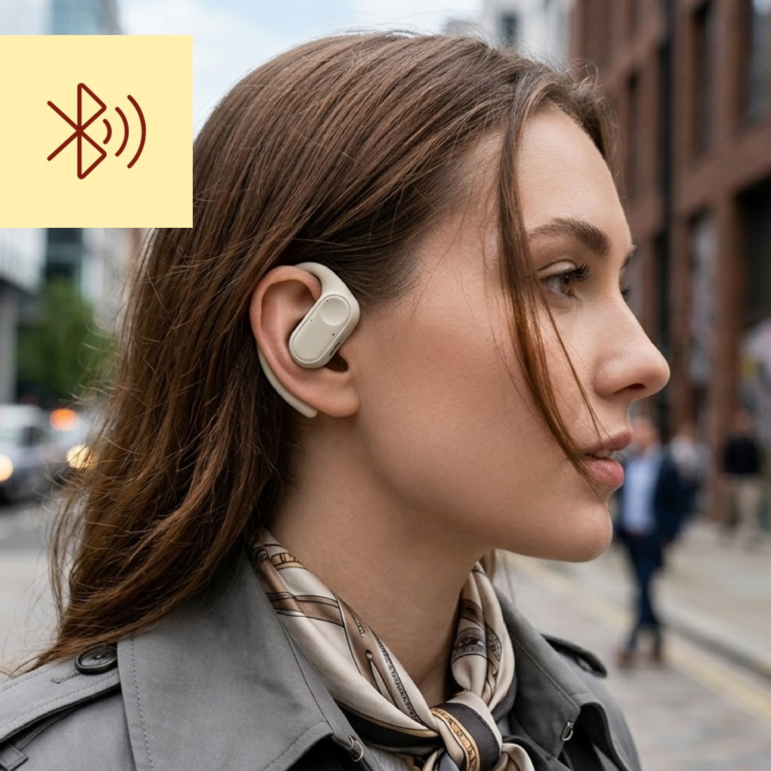 Eine Frau mit einem Bluetooth-Ohrhörer im rechten Ohr, die einen grauen Mantel und einen gemusterten Schal trägt, auf einer Stadtstraße.