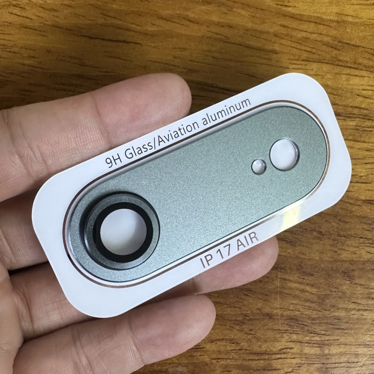 Una mano tiene una protezione per obiettivo della fotocamera argentata con testo '9H Glass/Aviation aluminum'.
