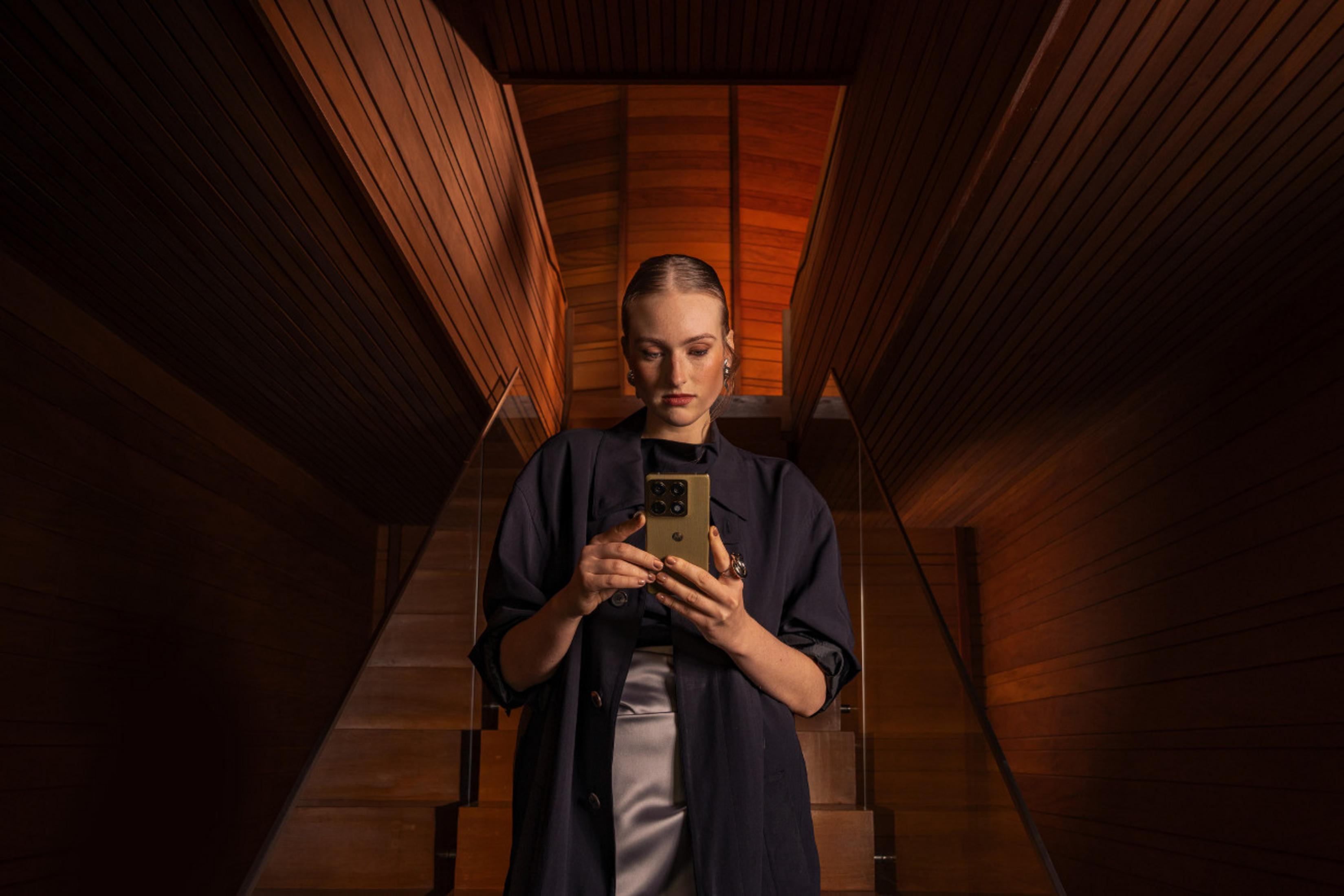Una donna in cappotto e gonna scuri è su scale di legno, guarda un telefono verde che tiene con entrambe le mani.