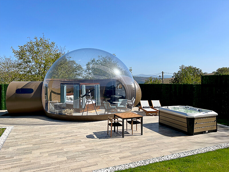 Una burbuja transparente con cama y telescopio, junto a un jacuzzi y mesa.