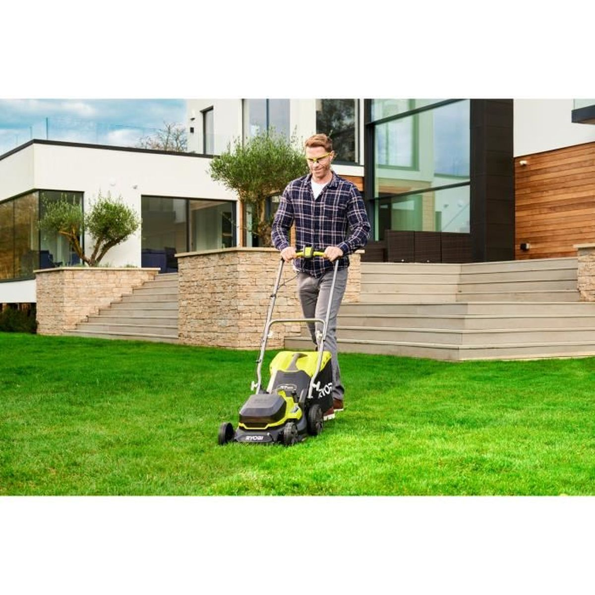 Mann mäht Rasen mit grünem Rasenmäher vor modernem Haus mit Steintreppen.