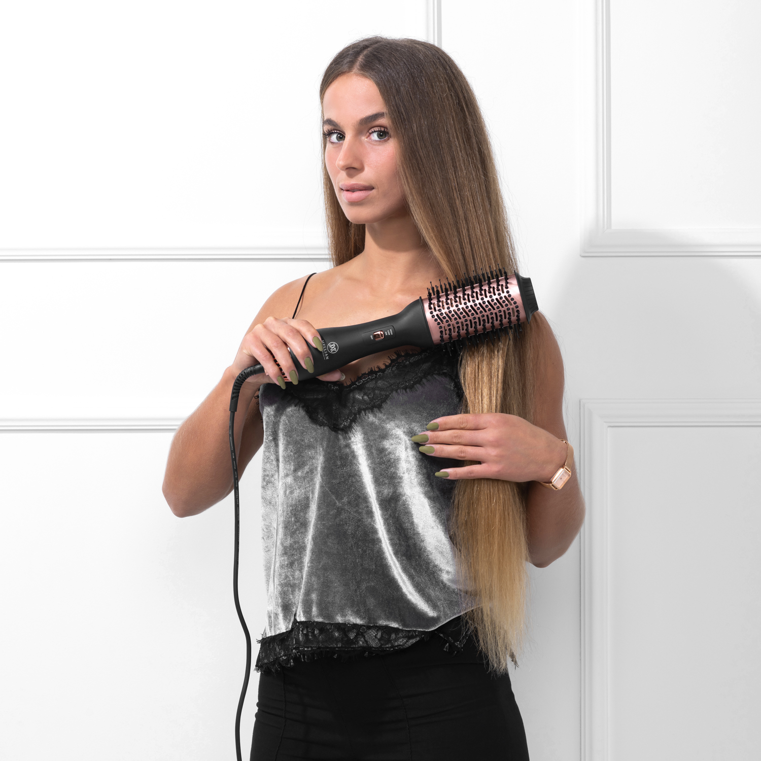 Eine Frau mit langen Haaren stylt ihr Haar mit einem roségoldenen Lockenstab vor einer weißen Wand.