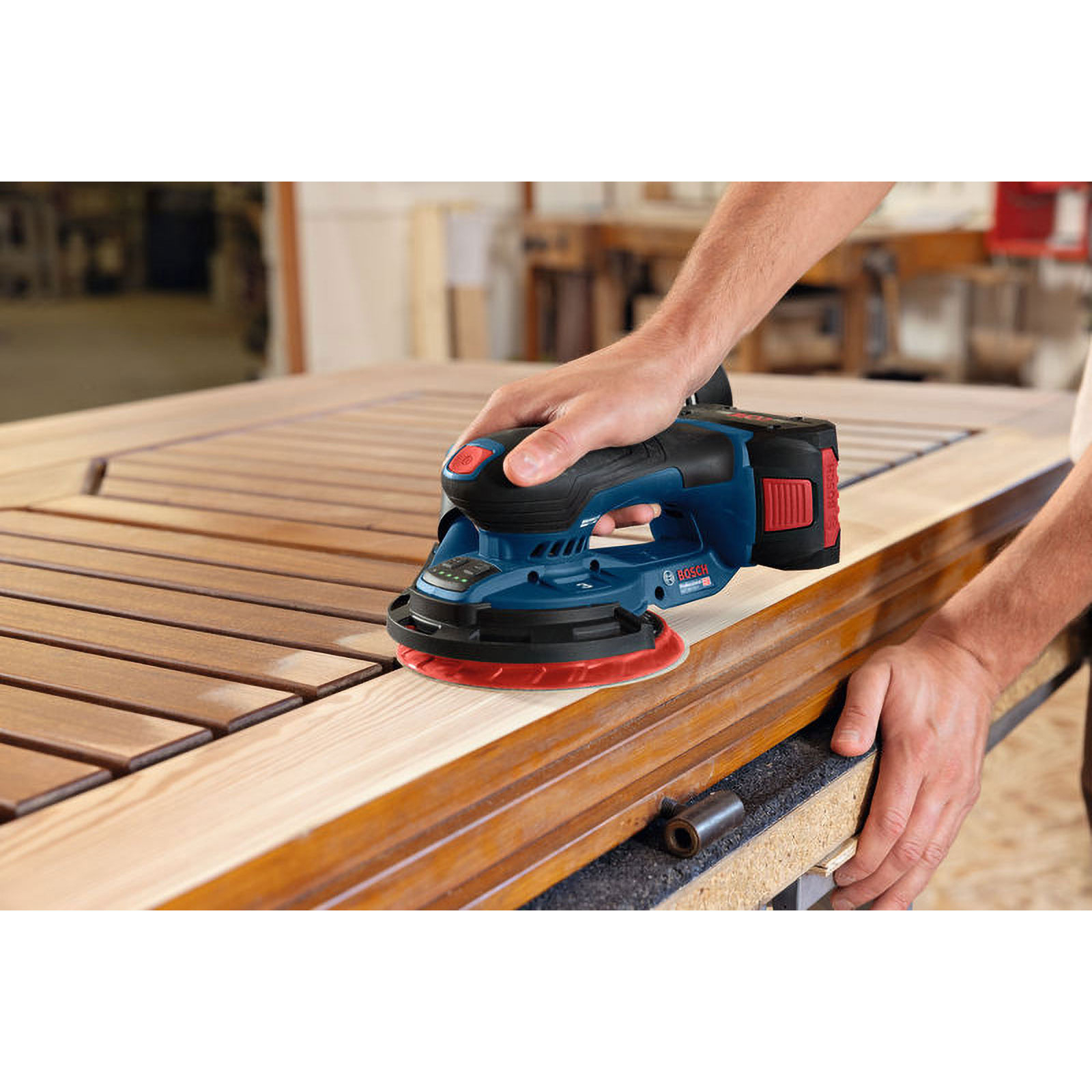 Ein Mann schleift Holz mit einem blauen Bosch-Schleifgerät in einer Werkstatt.
