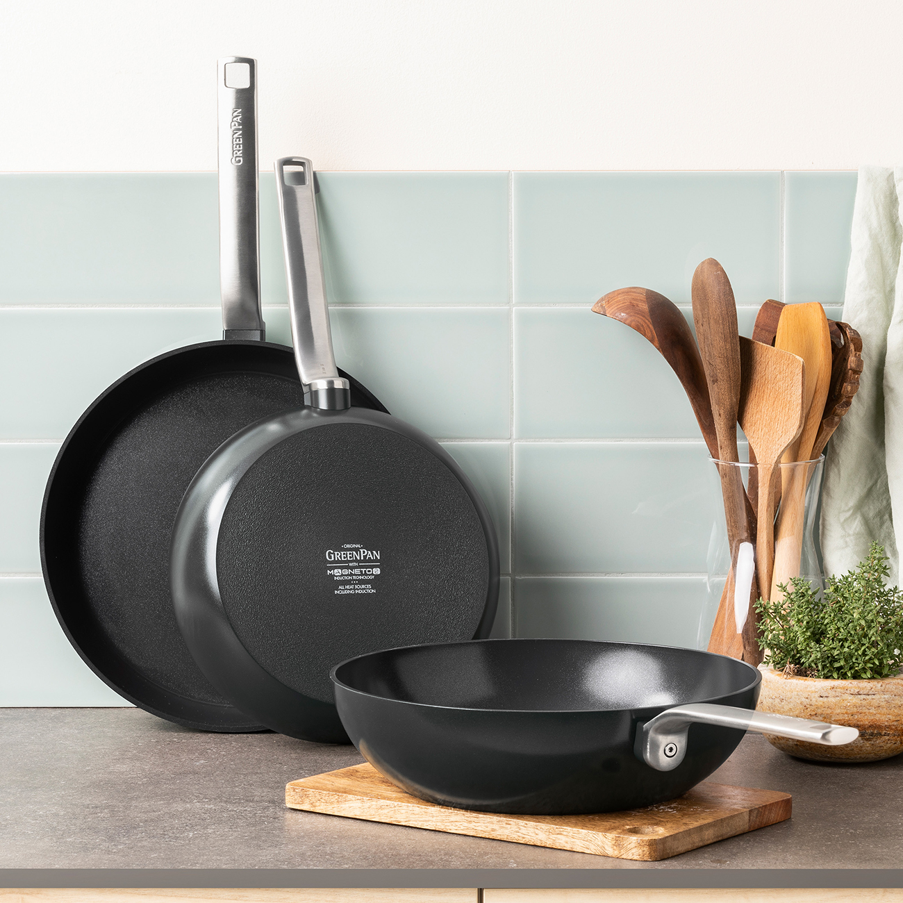 Drei schwarze Kochgeschirre, zwei Pfannen und ein Wok, auf einer Arbeitsplatte.