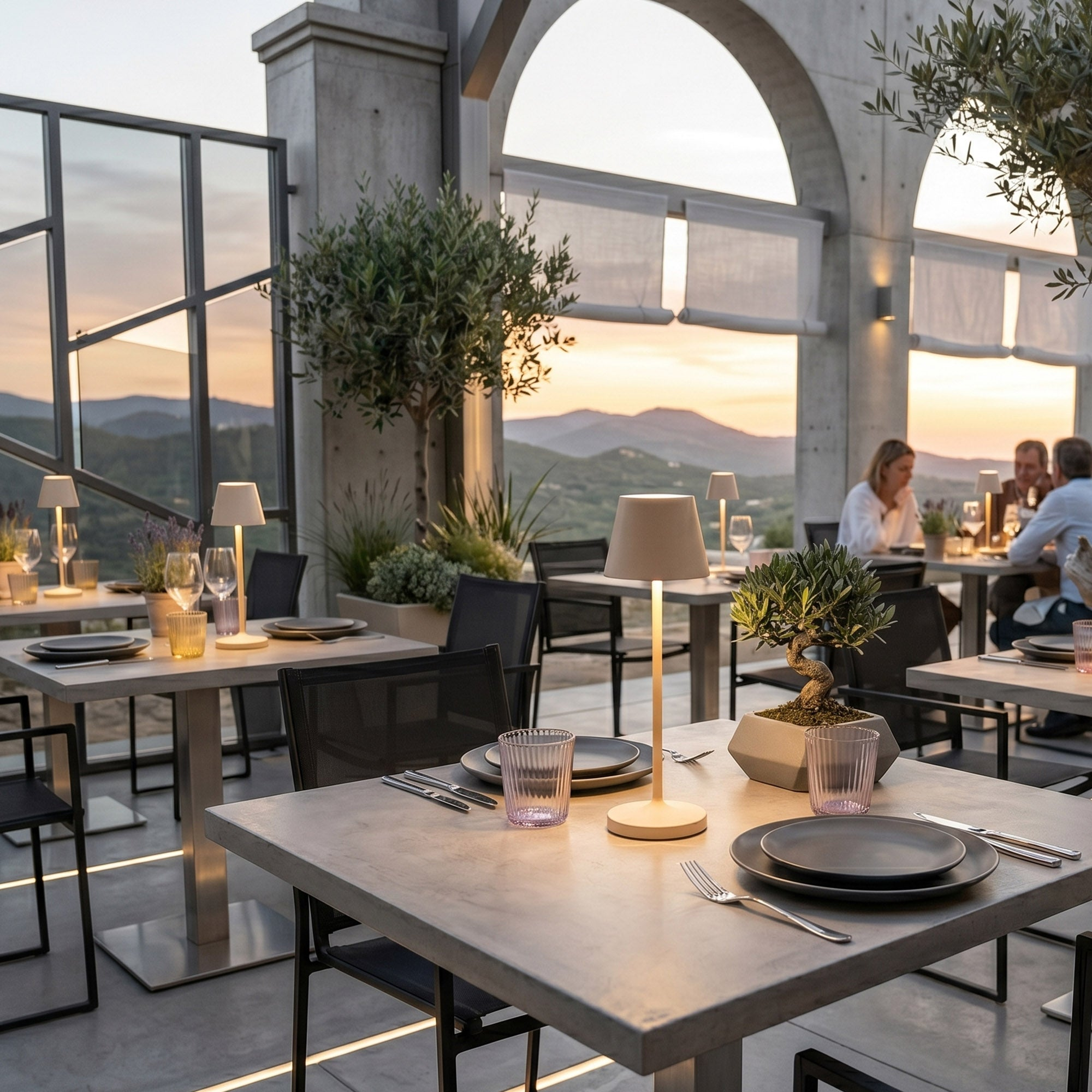 Tische mit Lampen und Gläsern auf einer Terrasse bei Sonnenuntergang mit Blick auf Hügel.