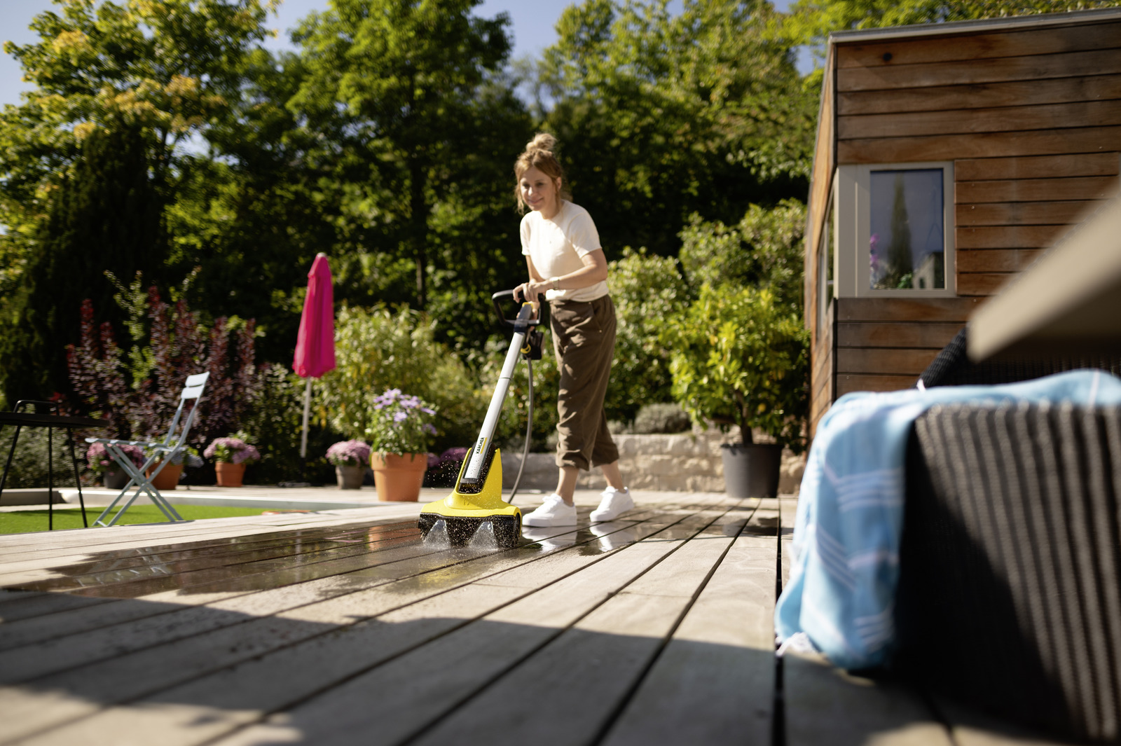 Frau reinigt Holzdeck mit elektrischer Bürste im Garten, sonnig.