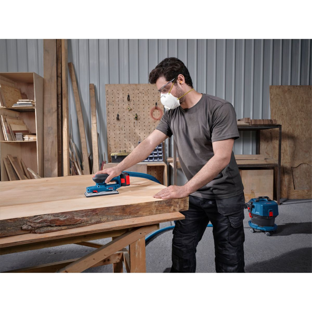 Mann schleift Holz mit blauem Schleifgerät, trägt Maske und Schutzbrille.