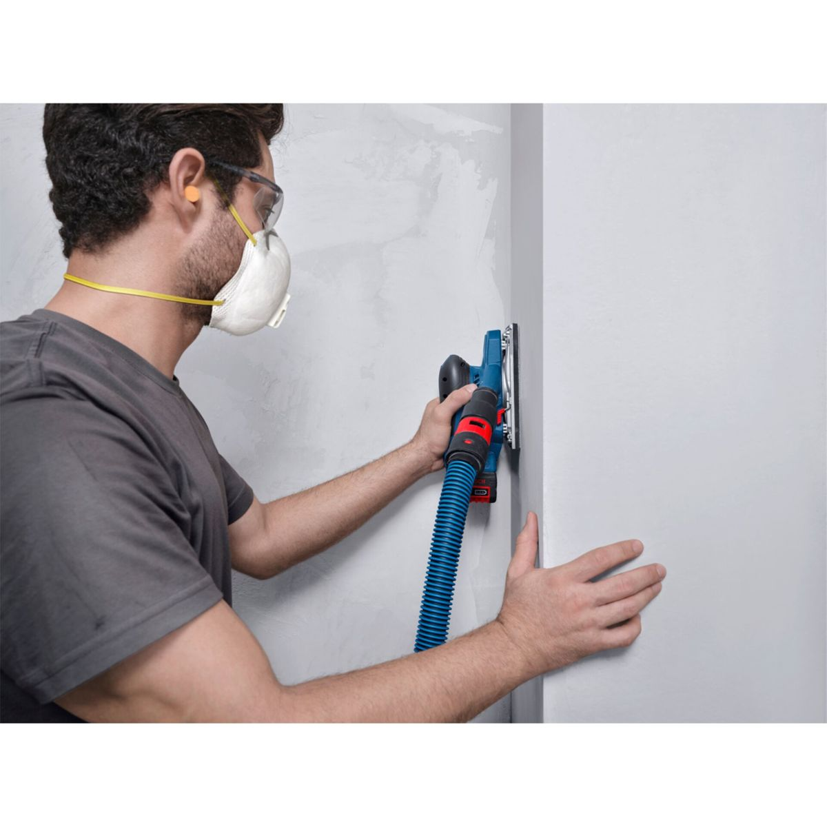Mann schleift Wand mit blauem Schleifgerät und Staubabsaugung.