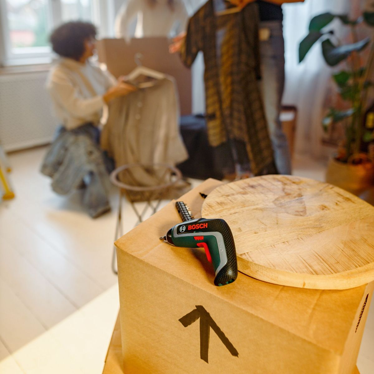 Ein kleiner Bosch-Akkuschrauber liegt auf Pappkartons, Menschen packen Kleidung aus.