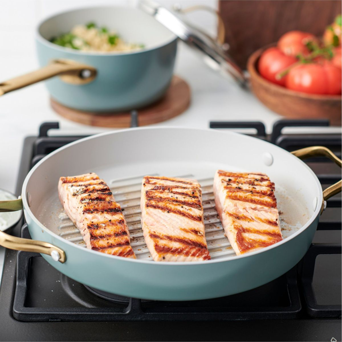 Drei gegrillte Lachsfilets in einer Pfanne mit Tomaten und einem Topf.