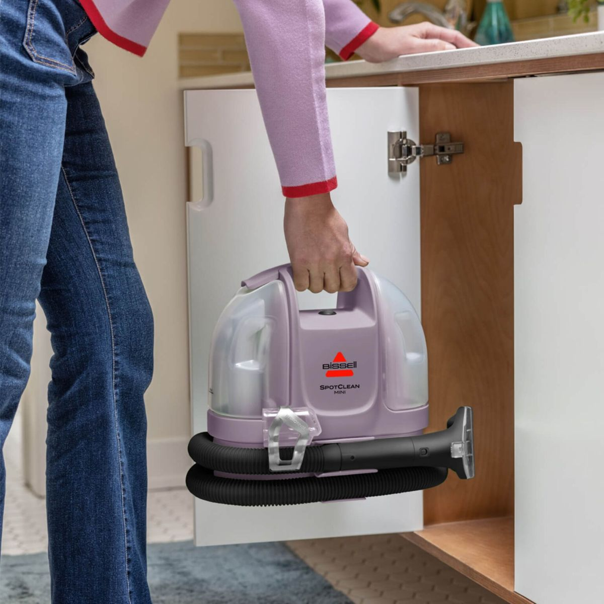 Eine Person mit Jeans hält einen lila Bissell SpotClean Mini Teppichreiniger.