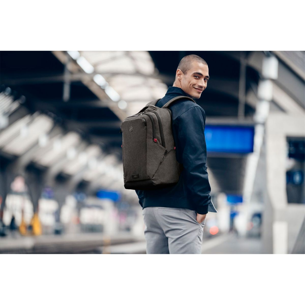 Mann mit Rucksack am Bahnhof, schaut zurück und lächelt.