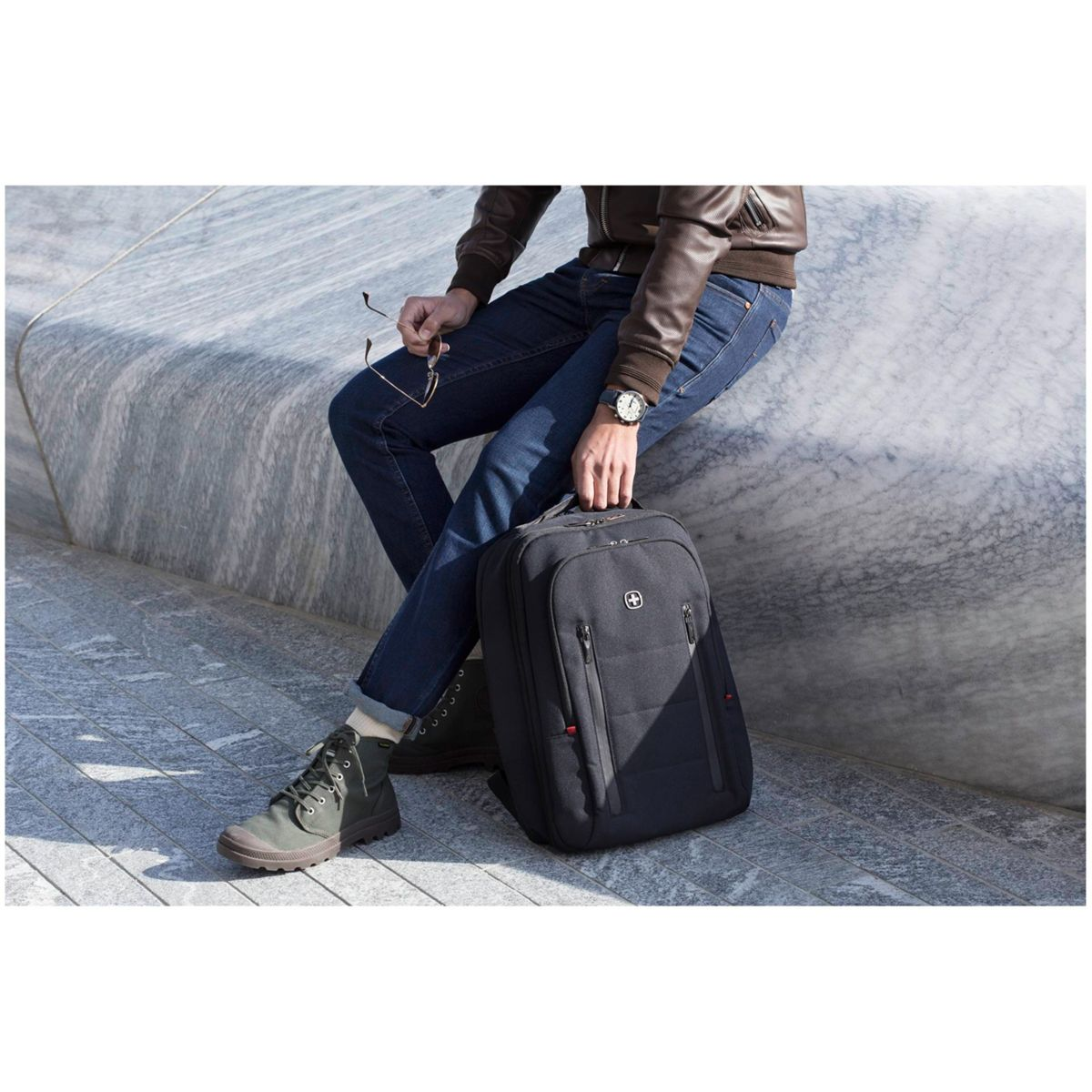 Mann in brauner Jacke, blauer Jeans und grünen Stiefeln sitzt mit schwarzem Rucksack.