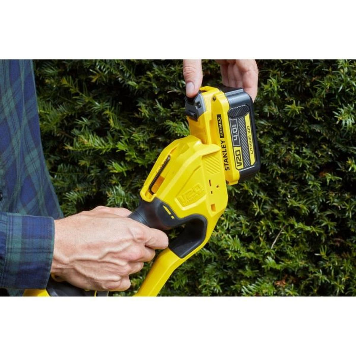 Una persona inserisce una batteria gialla in un attrezzo da giardino. Siepe verde sullo sfondo.