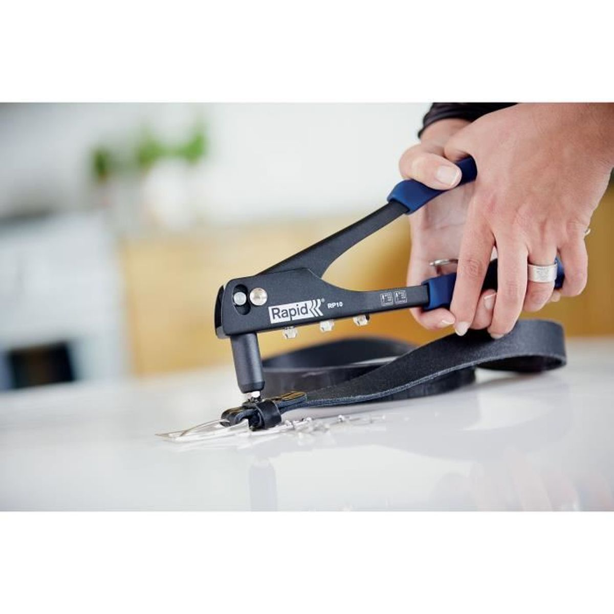 Una persona usa una rivettatrice Rapid RP10 nera con manici blu per perforare fori in una cinghia di cuoio nera su una superficie bianca.