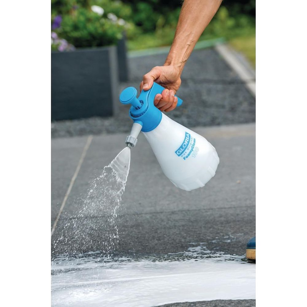 Una persona tiene in mano una bottiglia spray bianca e blu, spruzzando un liquido schiumoso bianco su una superficie pavimentata grigio scuro.