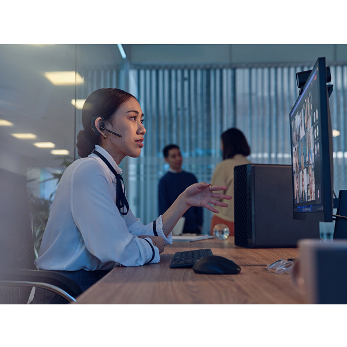 Donna con auricolare in videochiamata, due persone sullo sfondo, ufficio.