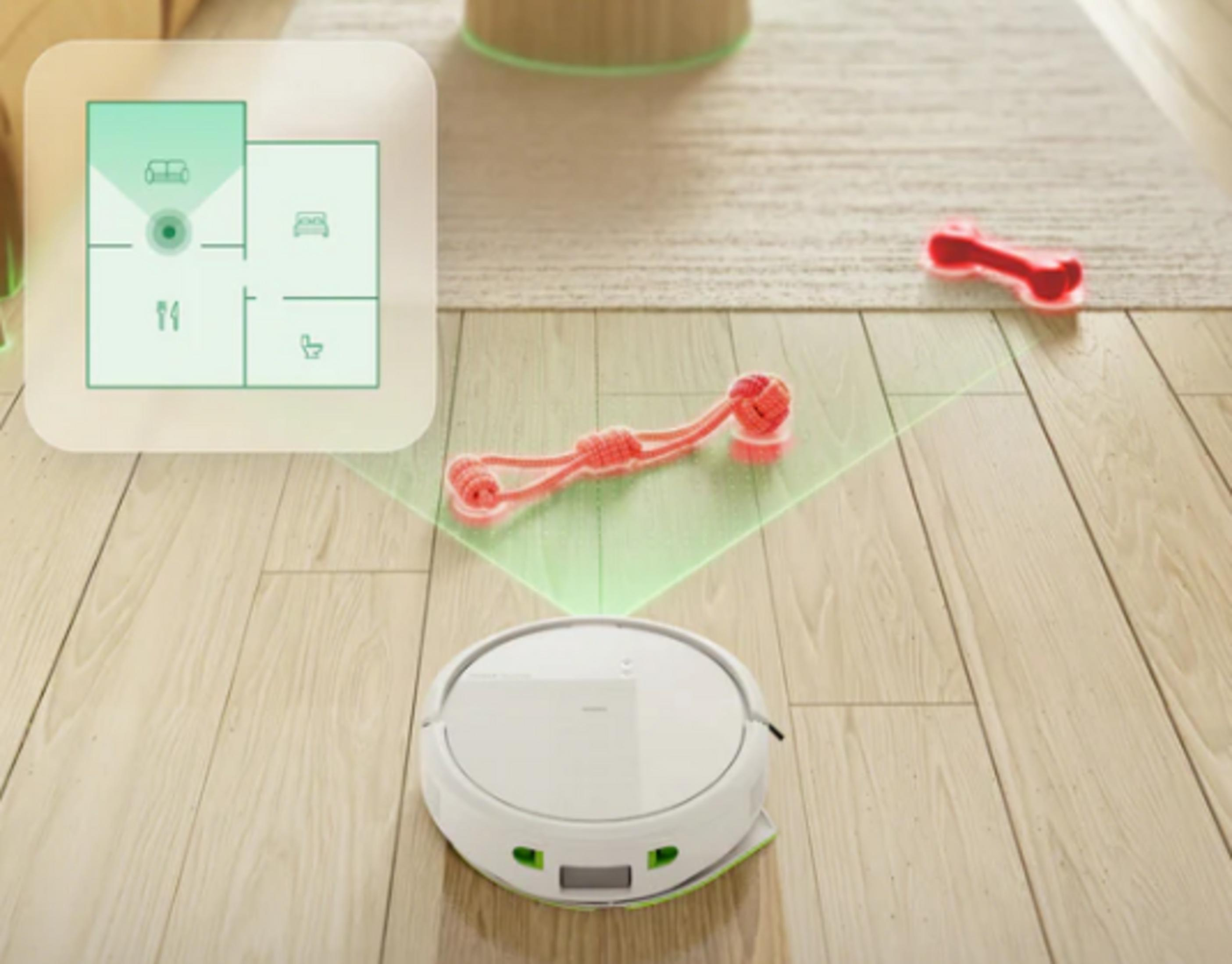Un robot aspirapolvere bianco pulisce un pavimento in legno vicino a un tappeto con giocattoli.