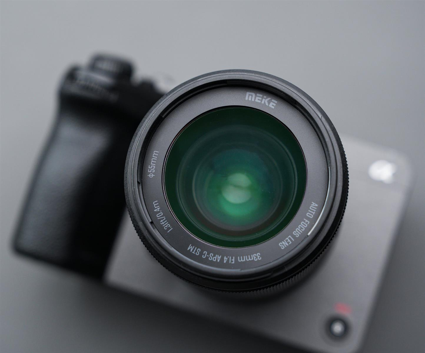 Ein schwarzes Kameraobjektiv, beschriftet mit „MEKE 33mm F1.4 APS-C STM AUTO FOCUS LENS“, auf einem grauen Kameragehäuse.
