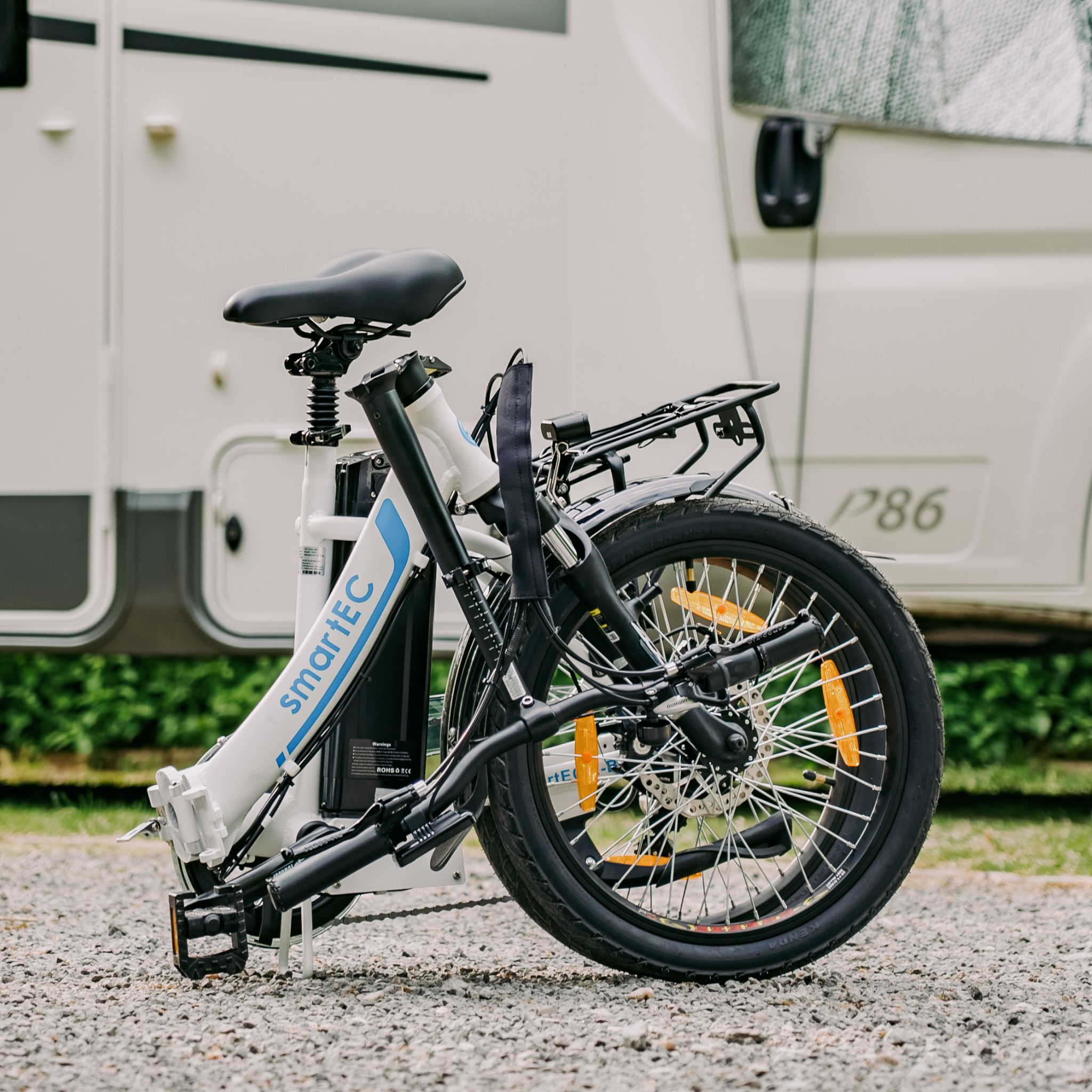 Ein weißes SmartEC E-Bike mit schwarzem Sattel und Gepäckträger steht auf Schotter vor einem Wohnmobil.