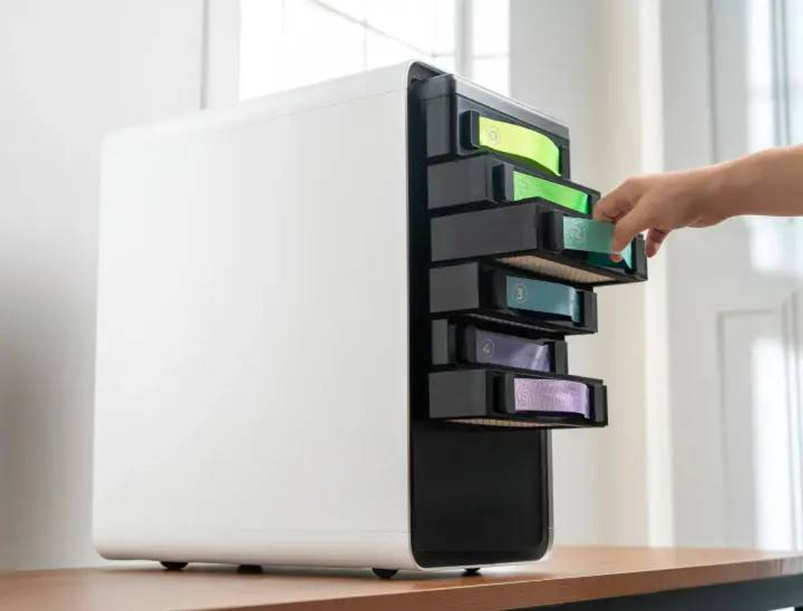 Una mano saca un cajón verde de un dispositivo blanco con varios cajones de colores.