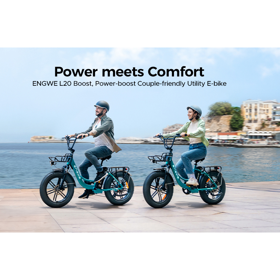 Ein Paar fährt türkisfarbene ENGWE L20 Boost E-Bikes entlang einer Uferpromenade mit Gebäuden und Meer im Hintergrund.
